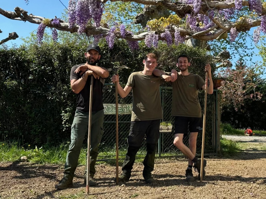 Équipe de jardiniers Etxelurra en pleine nature, posant avec leurs outils sous une glycine en fleurs après des travaux de paysagisme.